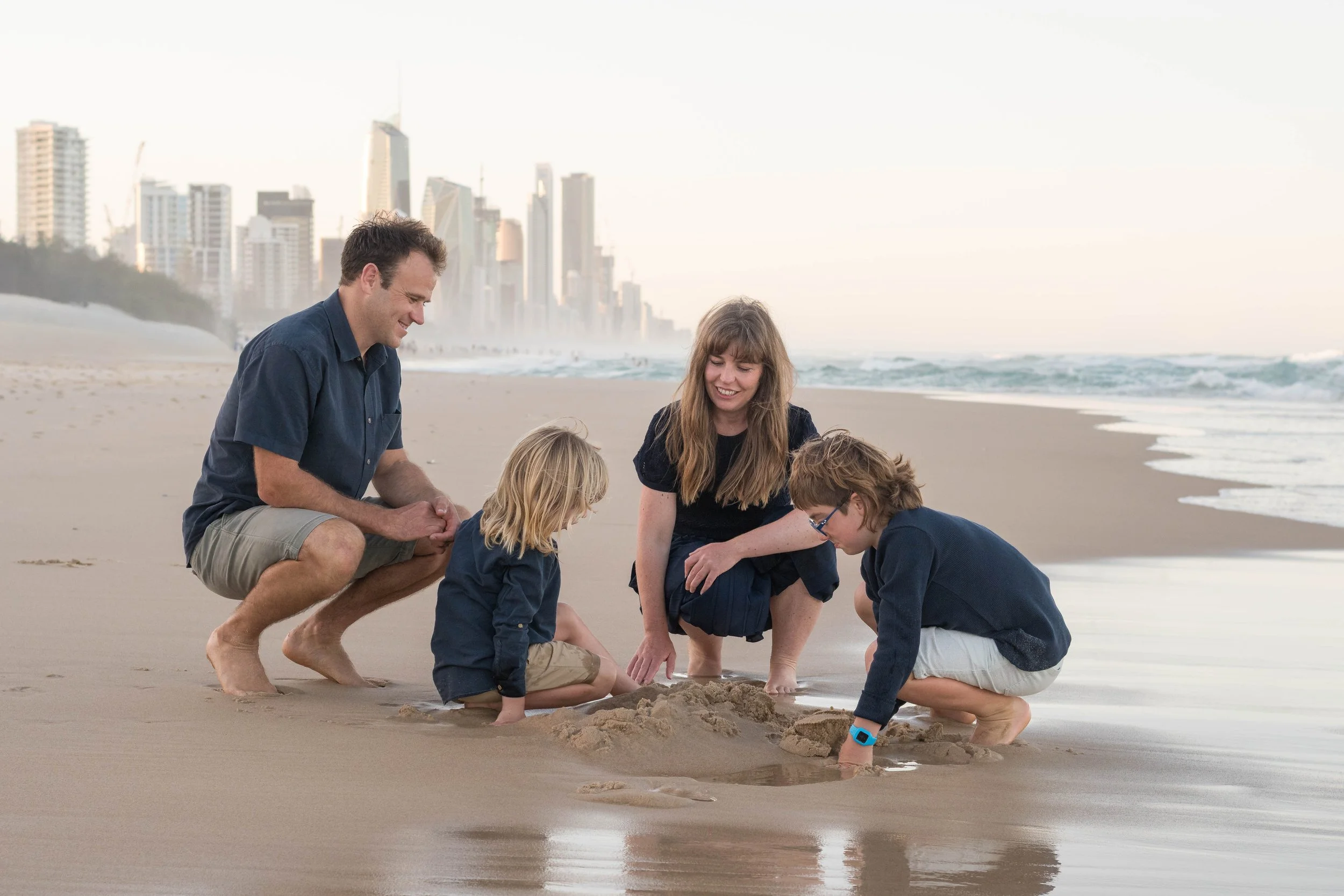 Family of four playing in the sand together with the city skyline behind them during a Mermaid Beach family portrait session.