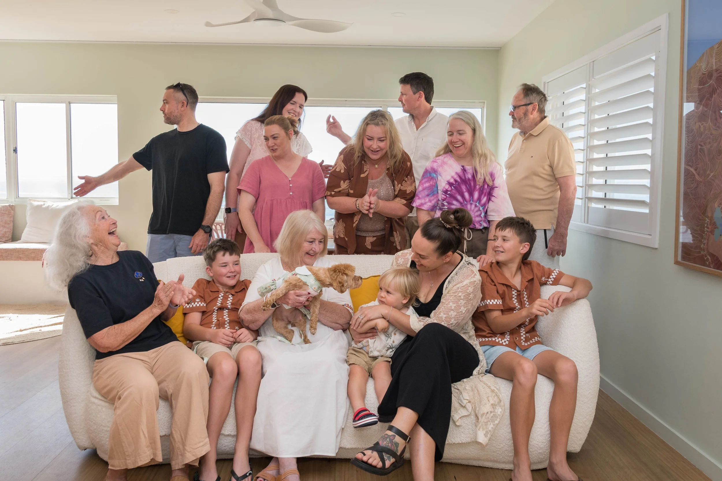 A large extended family across three generations gathered together on a sofa in a beach house living room at Broadbeach, wearing coordinated brown and neutral tones.