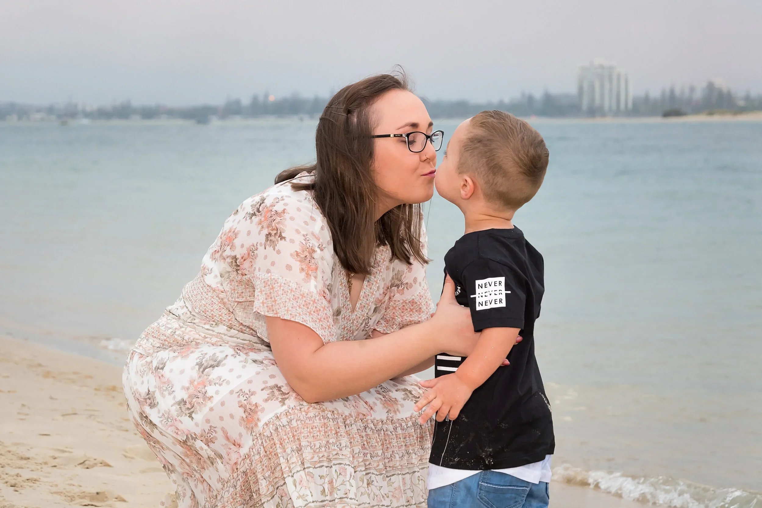 A mother in a floral dress kneeling to kiss her young son on the cheek at The Spit, with the Gold Coast skyline visible across the water.
