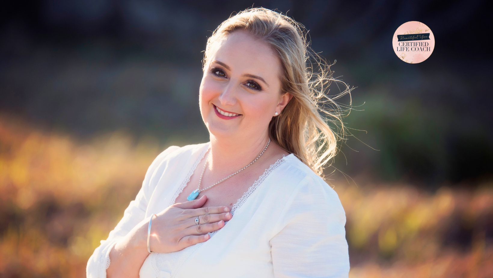 A smiling woman with blonde hair wearing a white top, a necklace with a blue pendant, and a ring, outdoors with sunlight and a blurred natural background. A pink circular badge with black and white text in the top right corner reads "Certified Life Coach."