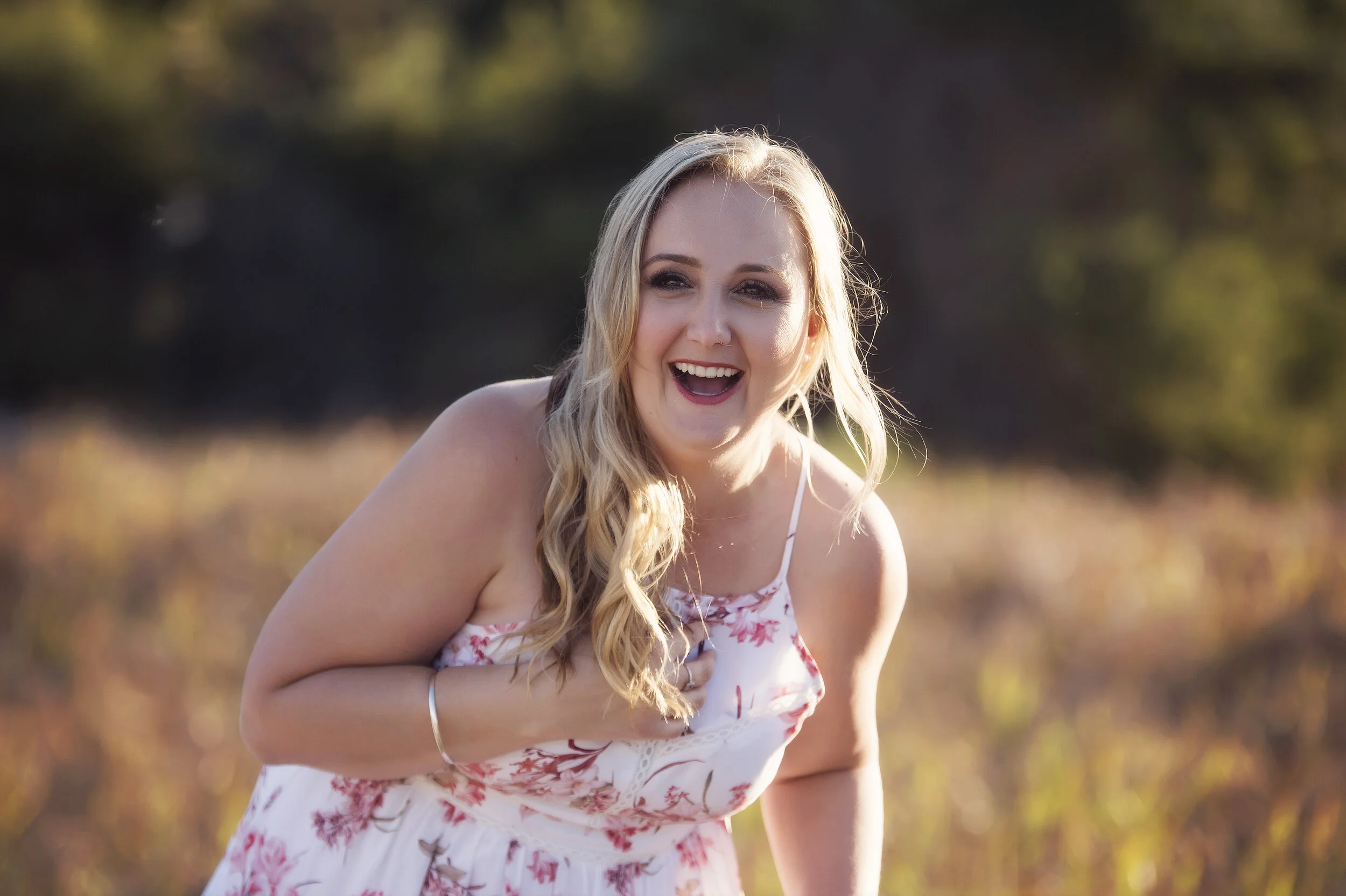 A woman with long blonde hair, wearing a floral dress, smiling and laughing outdoors on a sunny day.