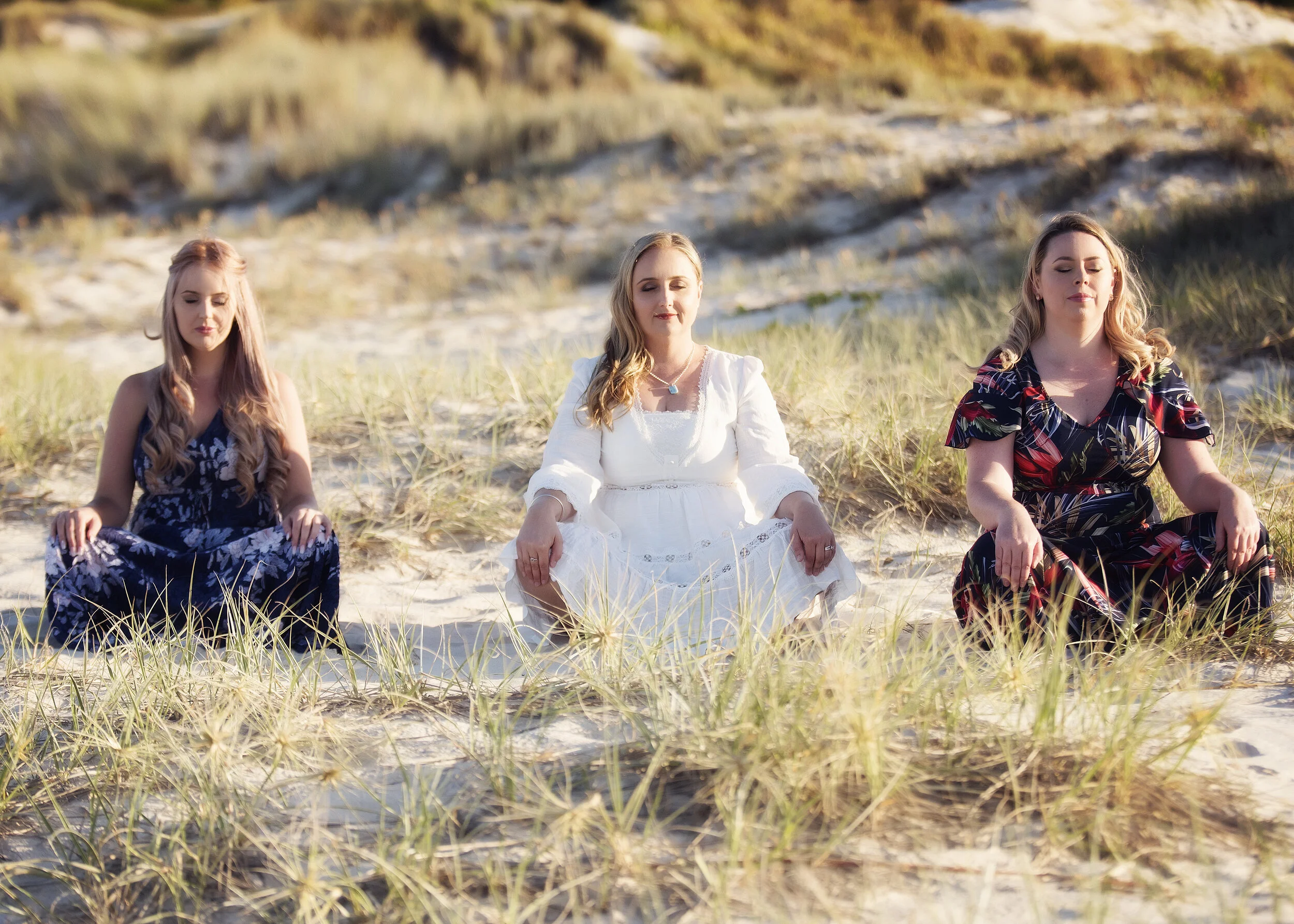 Three women sitting cross-legged on a sandy beach with dune grass, meditating with eyes closed, during sunset.