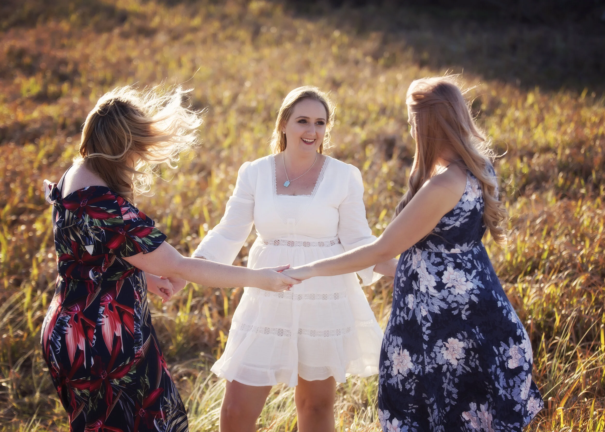 Three women in floral dresses holding hands and dancing in a field during sunset.