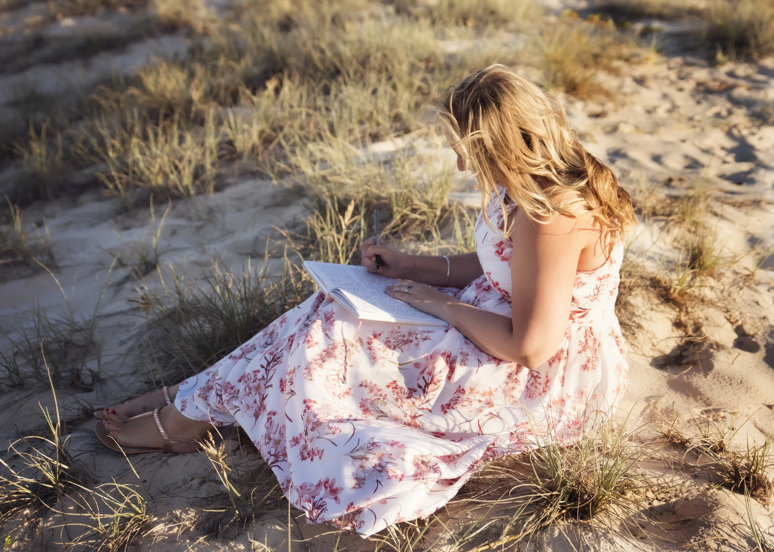 A woman with blonde hair wearing a floral dress sitting on sandy ground with grass, writing in a notebook with a pen.