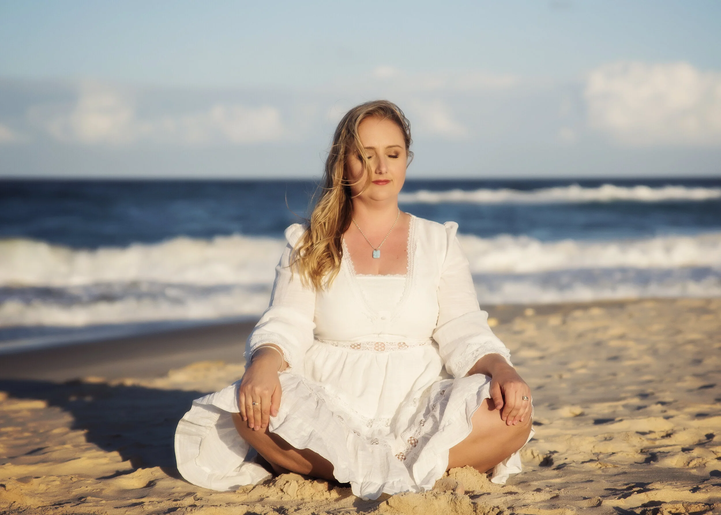 Woman meditating on the beach with ocean waves and blue sky in the background.