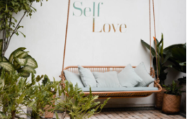 Bohemian-style hanging wicker sofa with white cushions, surrounded by lush green plants, and a wall with the words 'Self Love' in pastel colors.