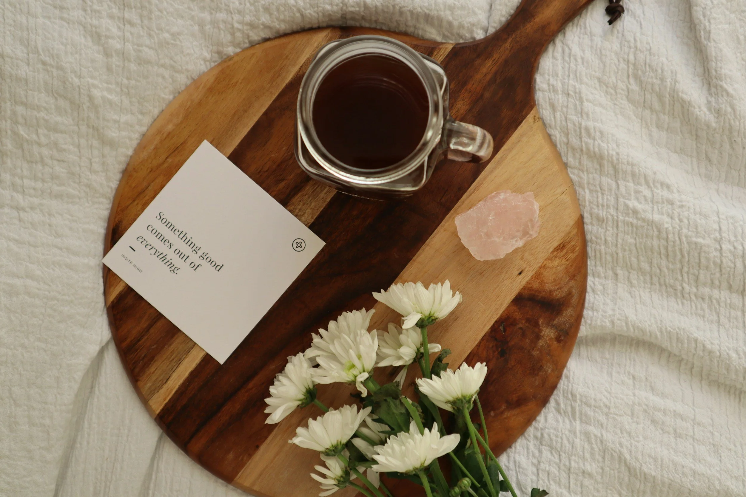 A round wooden serving board with a glass jar of dark liquid, a piece of pink salt, a small bouquet of white daisies, and a card with the quote 'Something good comes out of everything.' The board is on a white textured bedspread.