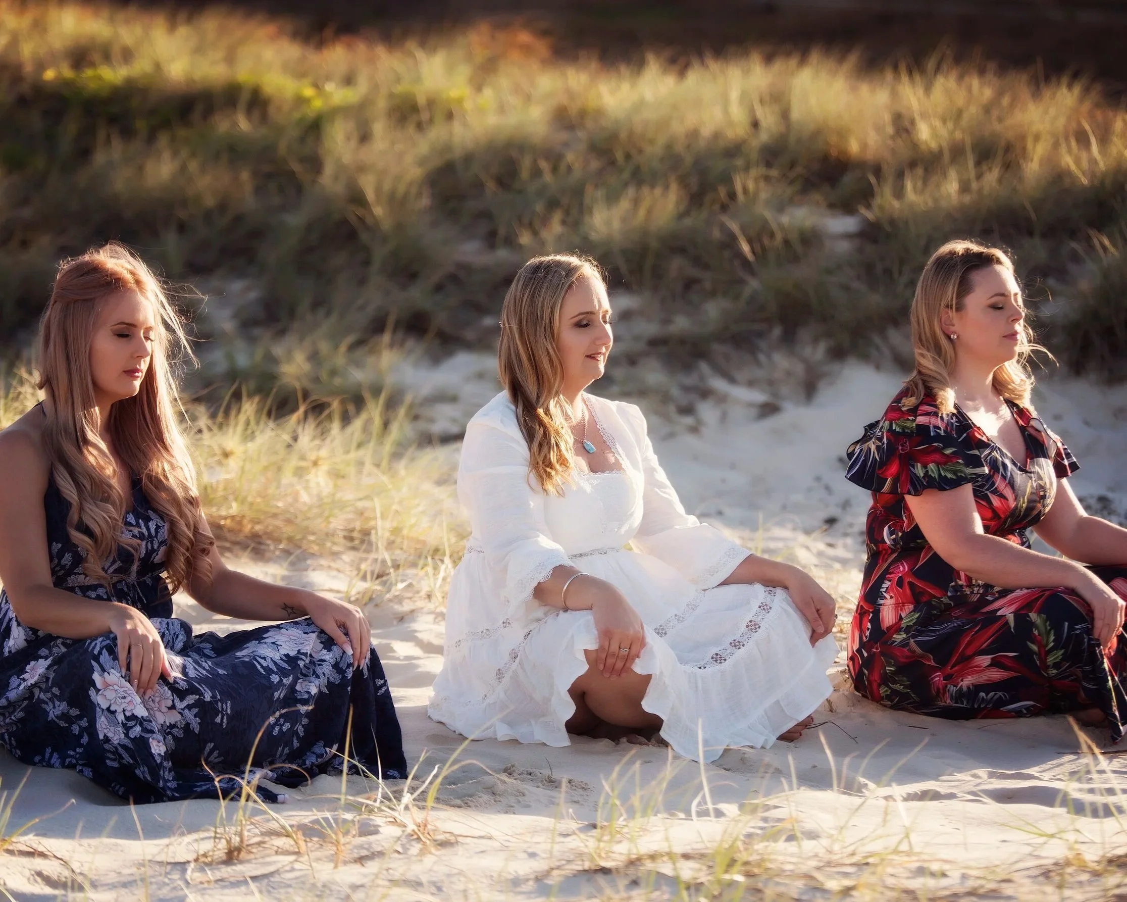 Three women sitting cross-legged on the sand in a meditative pose at the beach with grass-covered dunes in the background, wearing floral dresses.