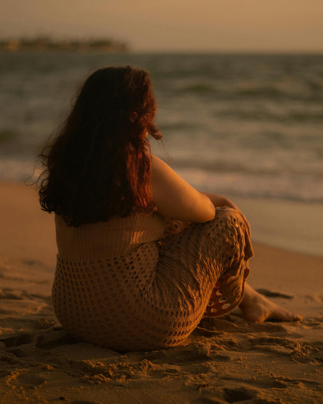 A woman with long, dark hair sitting alone on a sandy beach during sunset, gazing at the ocean.