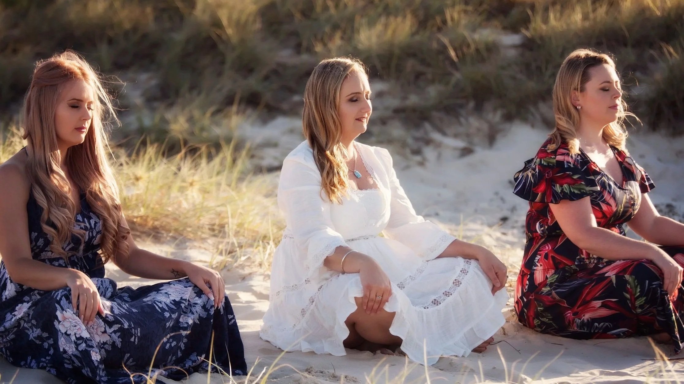 Three women sitting cross-legged on a sandy beach with grassy dunes in the background, eyes closed and meditating.