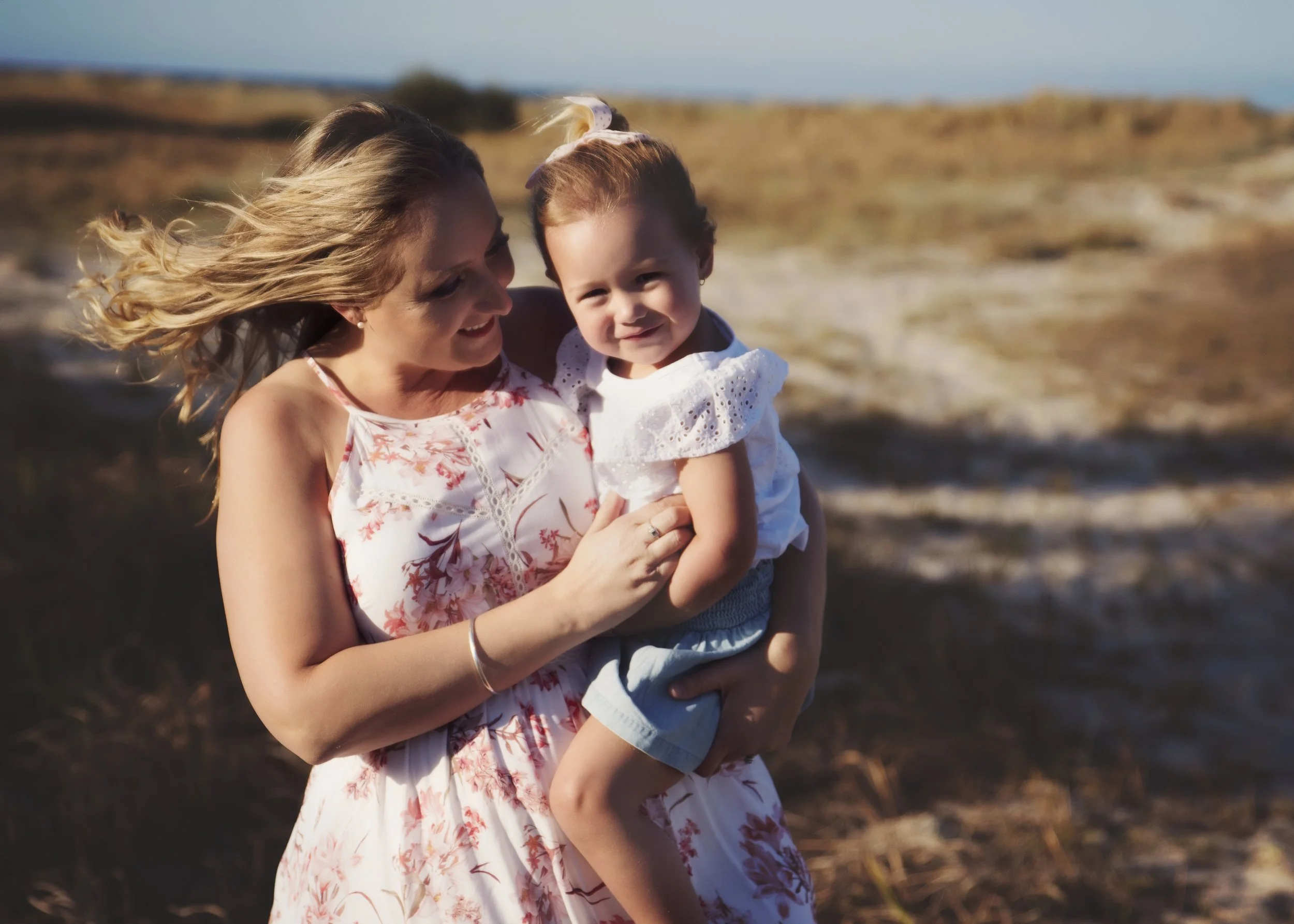 A woman holding a young girl outdoors near a beach or fields, both smiling.