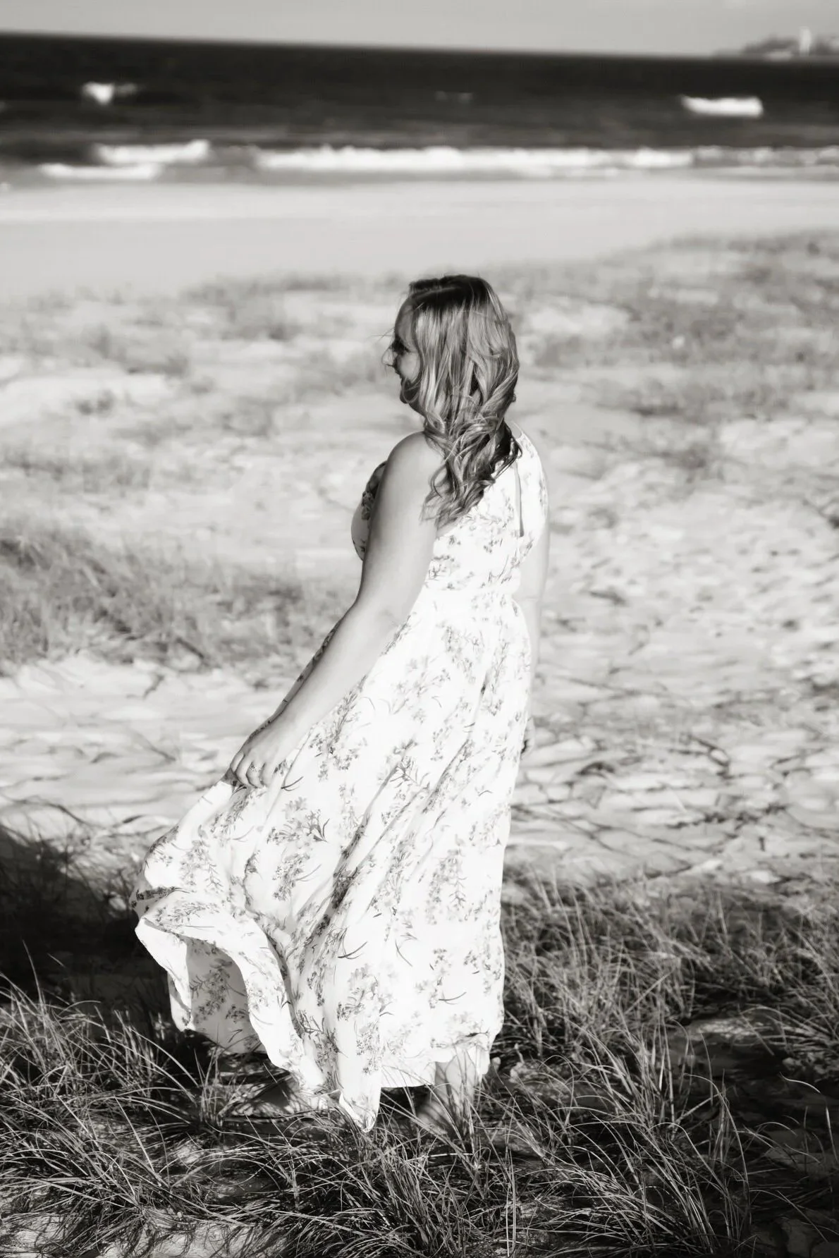 A woman in a floral maxi dress walking along a sandy beach with grass in the foreground and ocean waves in the background.