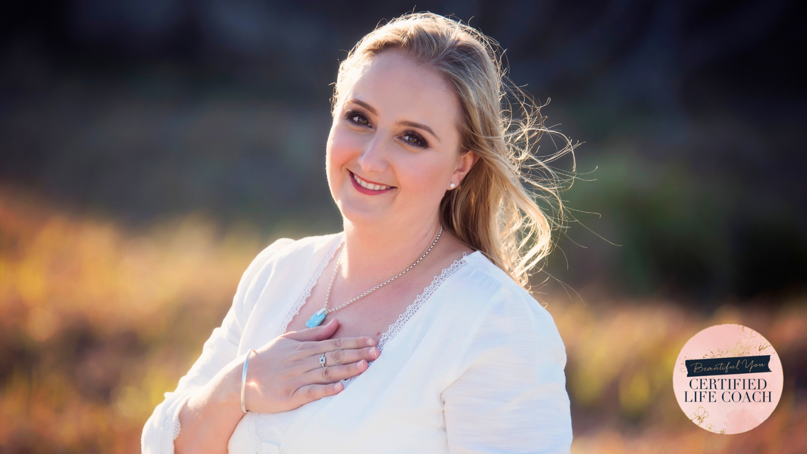A woman with blonde hair wearing a white top, smiling and holding her hand over her chest, outdoors during golden hour with blurred background.