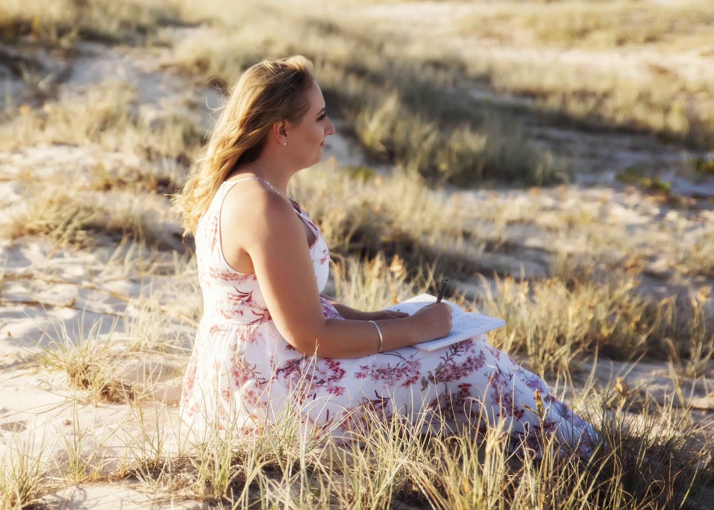 A woman with long blonde hair sitting on sandy grass at the beach, taking notes in a notebook with a pen.