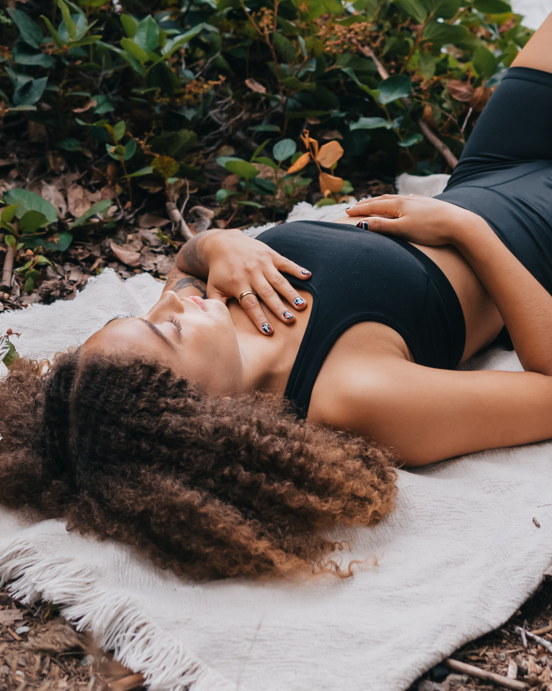 A woman with curly hair lying on a towel outdoors among leaves, resting with her hand on her chest.