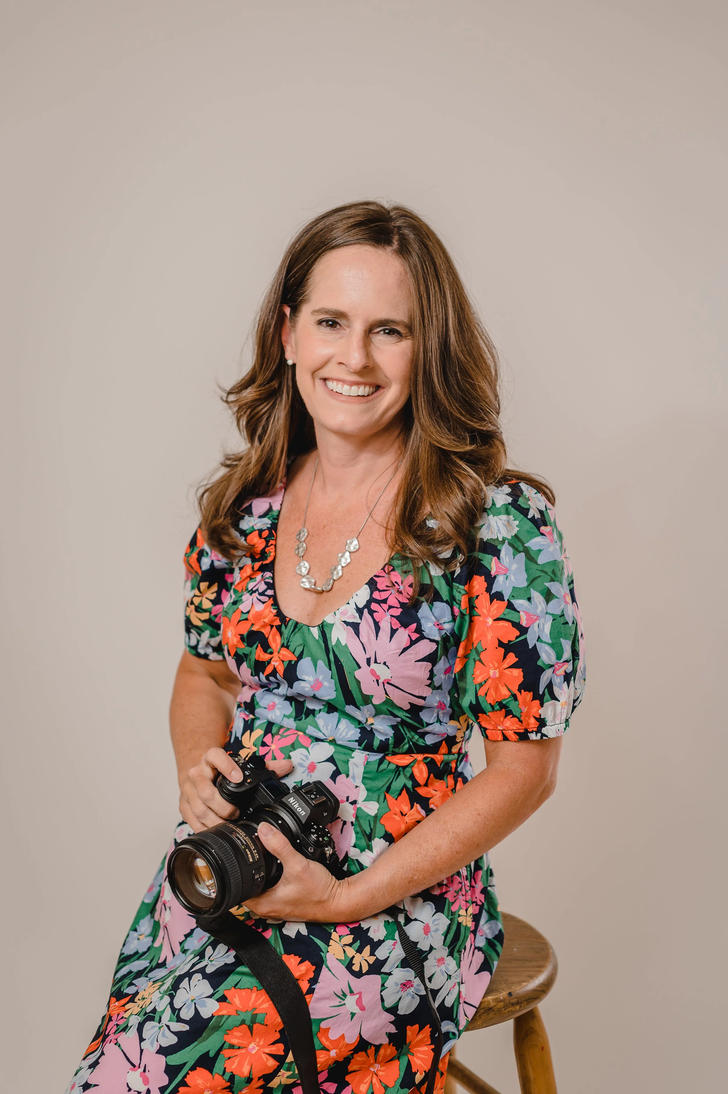 A photographer in a flowered dress sits on a stool holding her camera and smiling