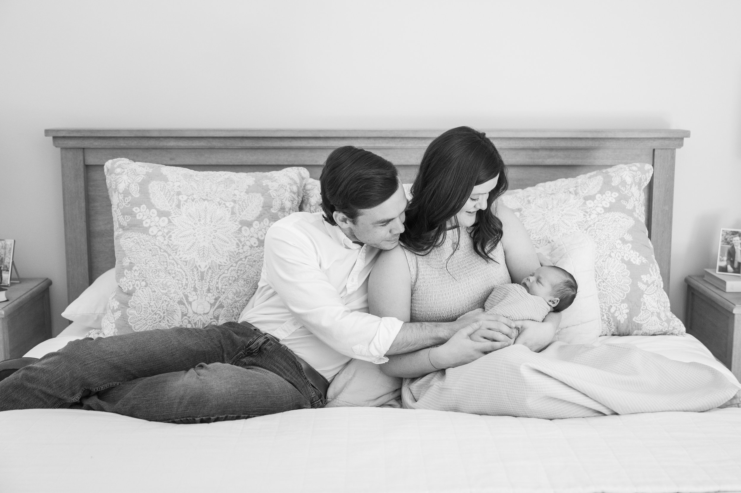 a black and white image of a new mom and dad sitting on a bed, smiling and staring at their sleeping newborn