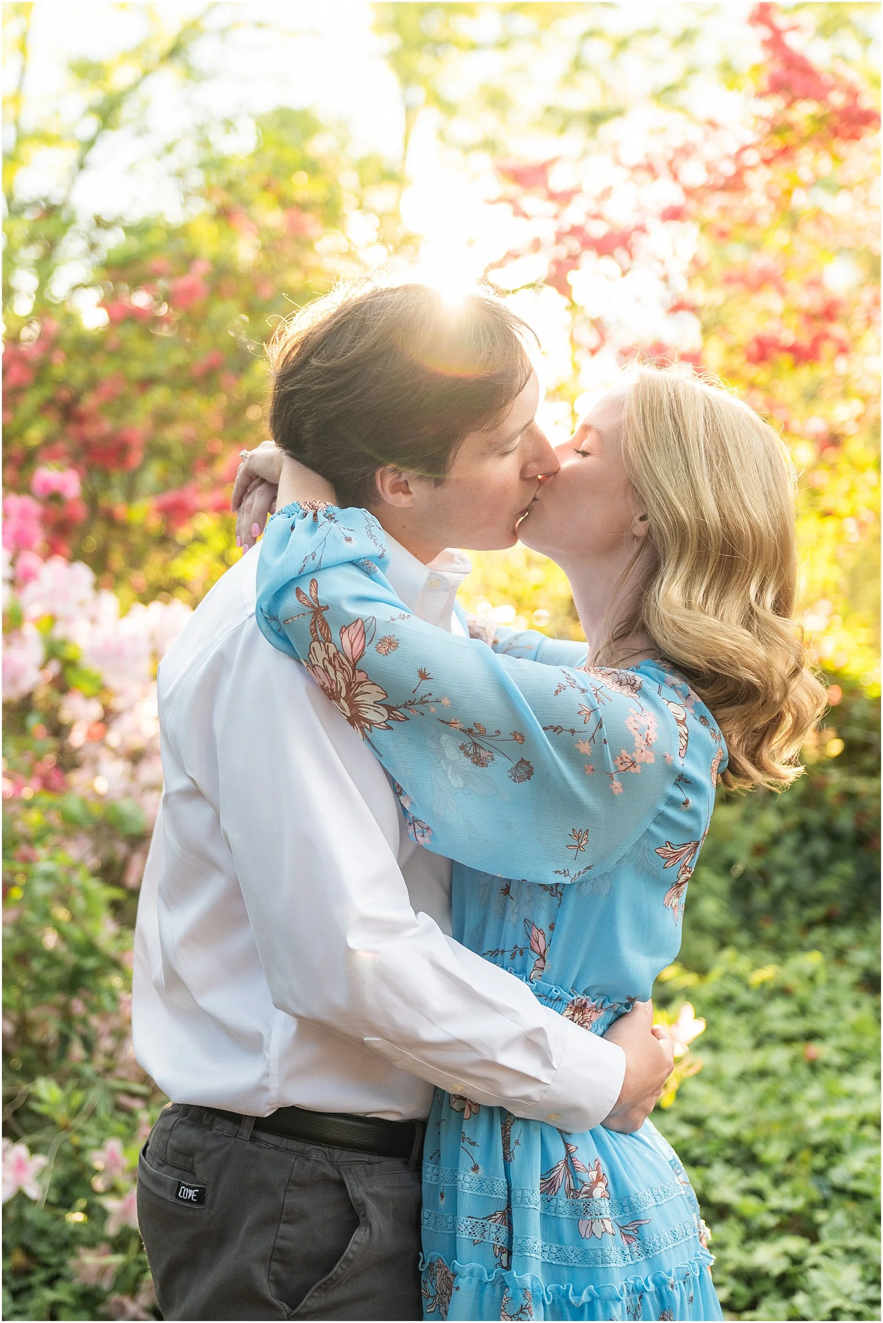 Young man and woman kissing as the sun sets behind them, surrounded by an azalea garden.