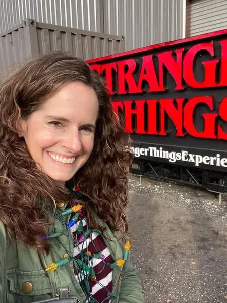 woman dressed as Joyce Byers smiling in front of the Stranger Things Experience sign
