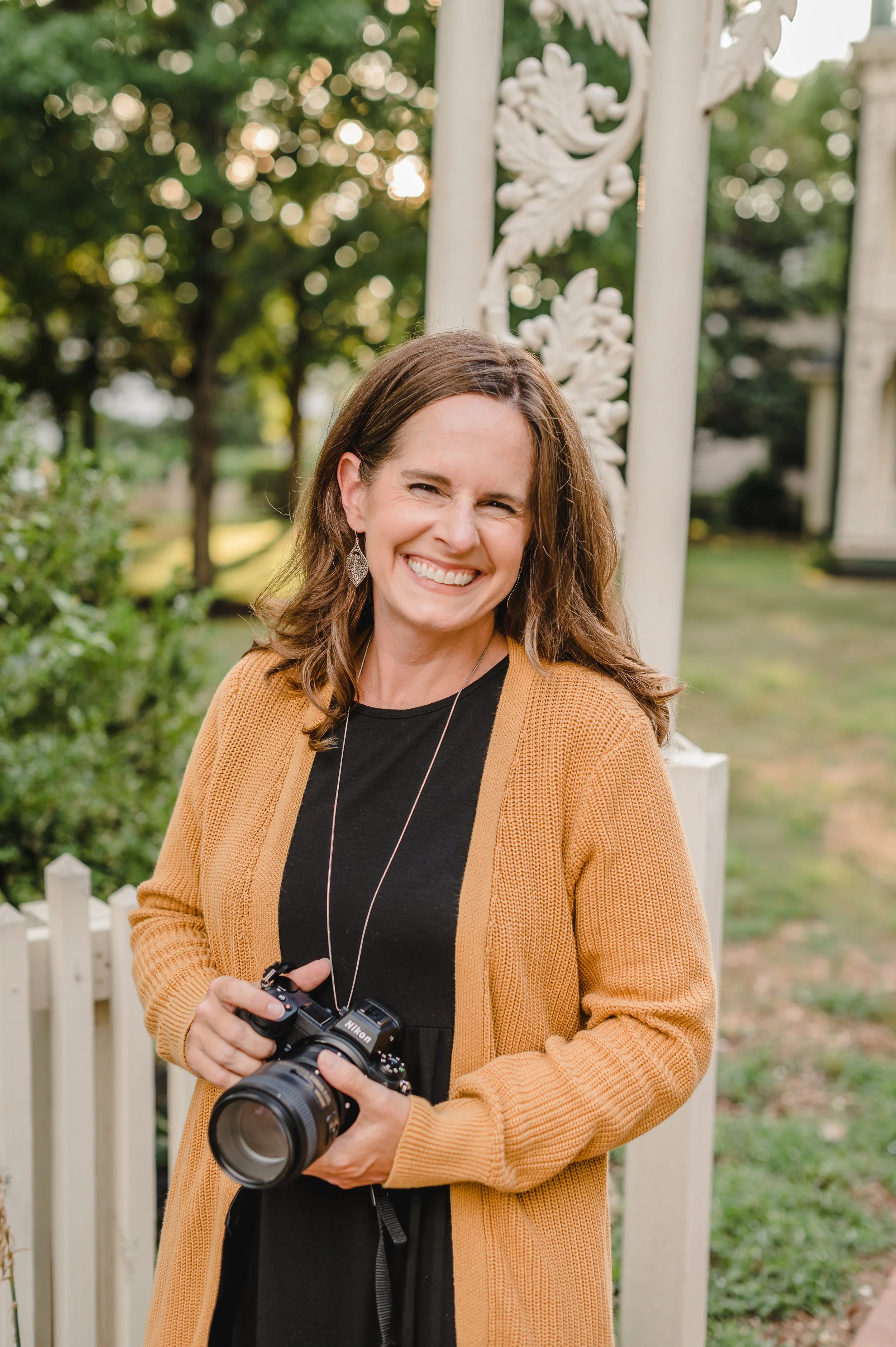 A photographer in a black top with a yellow sweater holds her camera and grins happily