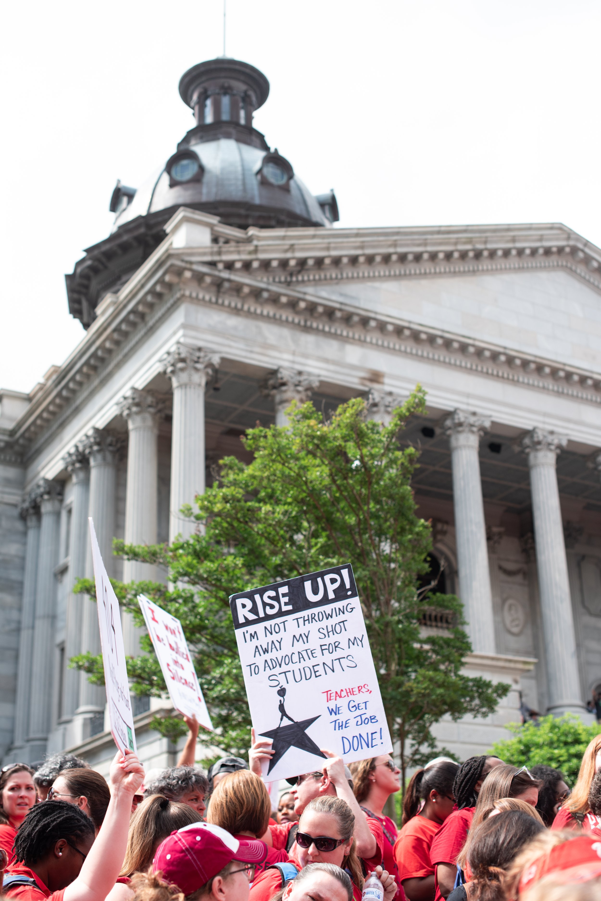 A group of teachers wearing red and holding signs stand in front of the SC State House in May of 2019, rallying for better public education.