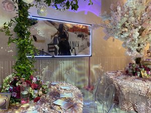 Elegant event setup with floral centerpieces, clear chairs, and a large framed poster on the wall. The tables are decorated with pink and purple flowers, alongside menu cards and candles in glass holders. A decorative tree with white blossoms adds a 