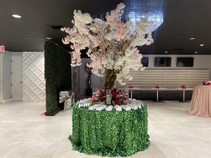 Elegant event setup with a table adorned with pink and white faux cherry blossoms, surrounded by name cards. Table covered with green foliage cloth and red roses in vases. Plush seating area in background with pink rose bouquets.