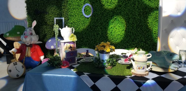 Alice in Wonderland themed table with a white rabbit statue holding a clock, a tea set on a blue cloth, potted yellow roses, artificial greenery, a faux hedge backdrop, mushroom decorations, and whimsical decor including a teapot and checkered cloth.