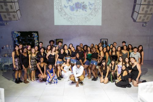 Group of young people posed in formal attire at an indoor event venue with floral decorations and a projected message reading "Have Courage and Be Kind."