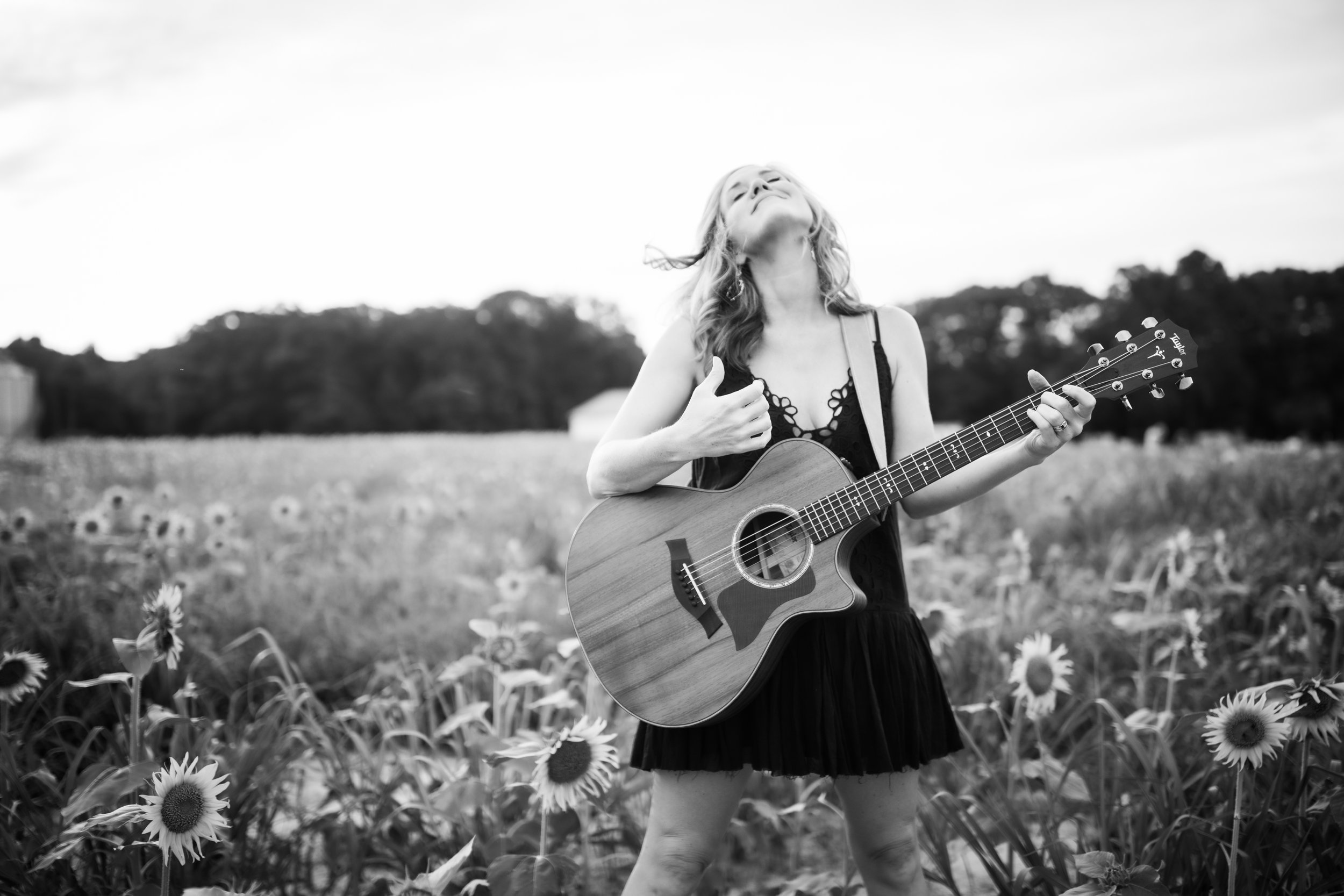 Black and white photo of a woman playing an acoustic guitar in a sunflower field.
