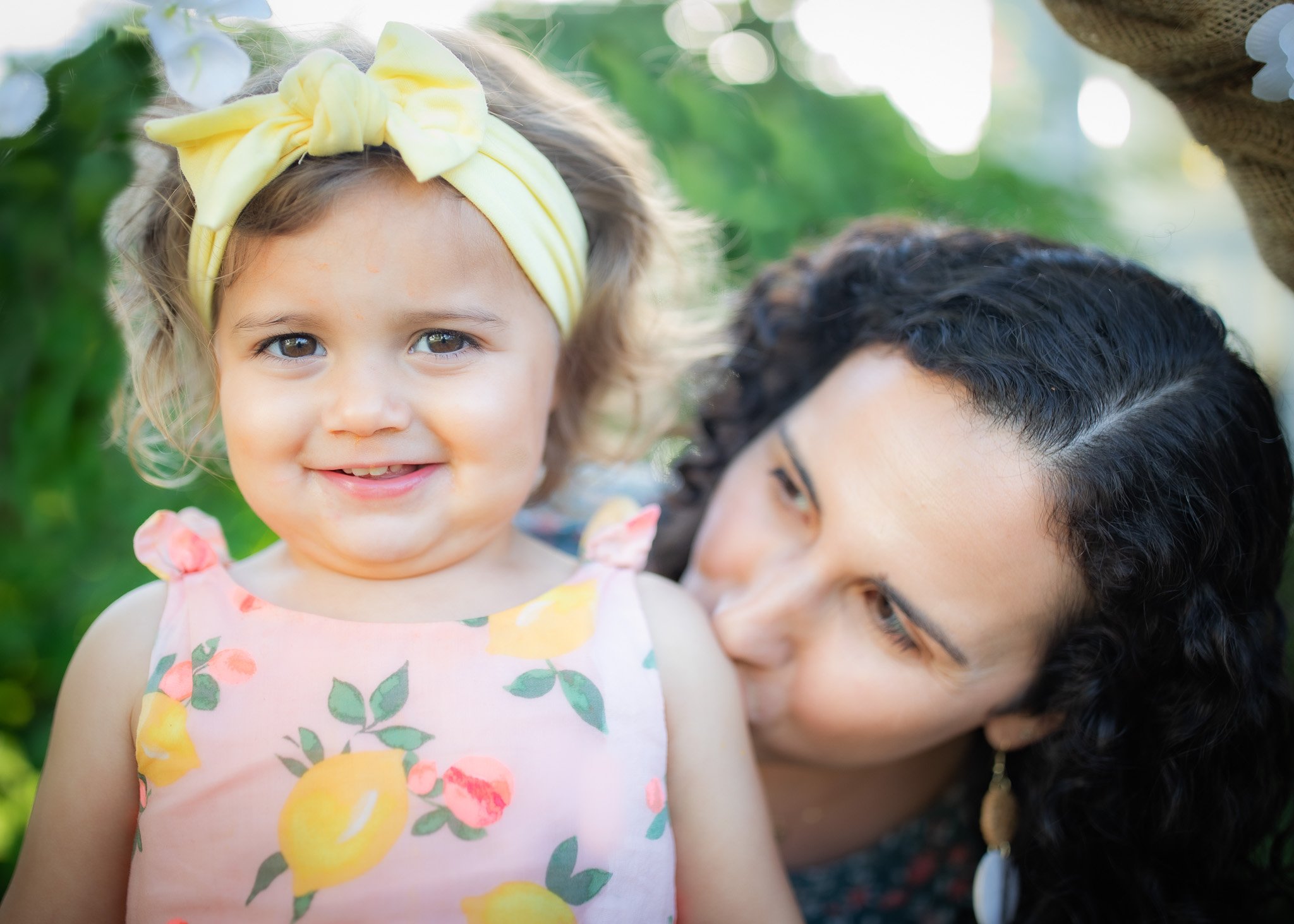 Smiling child with yellow bow headband and floral dress, adult woman kissing child's shoulder outdoors, green foliage background.