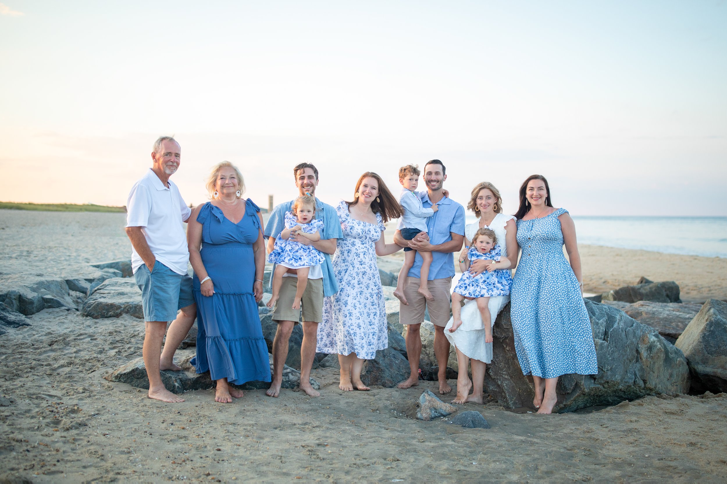 Family group portrait on a beach with adults and children, wearing blue and white clothing, standing on sand with rocks in the background.