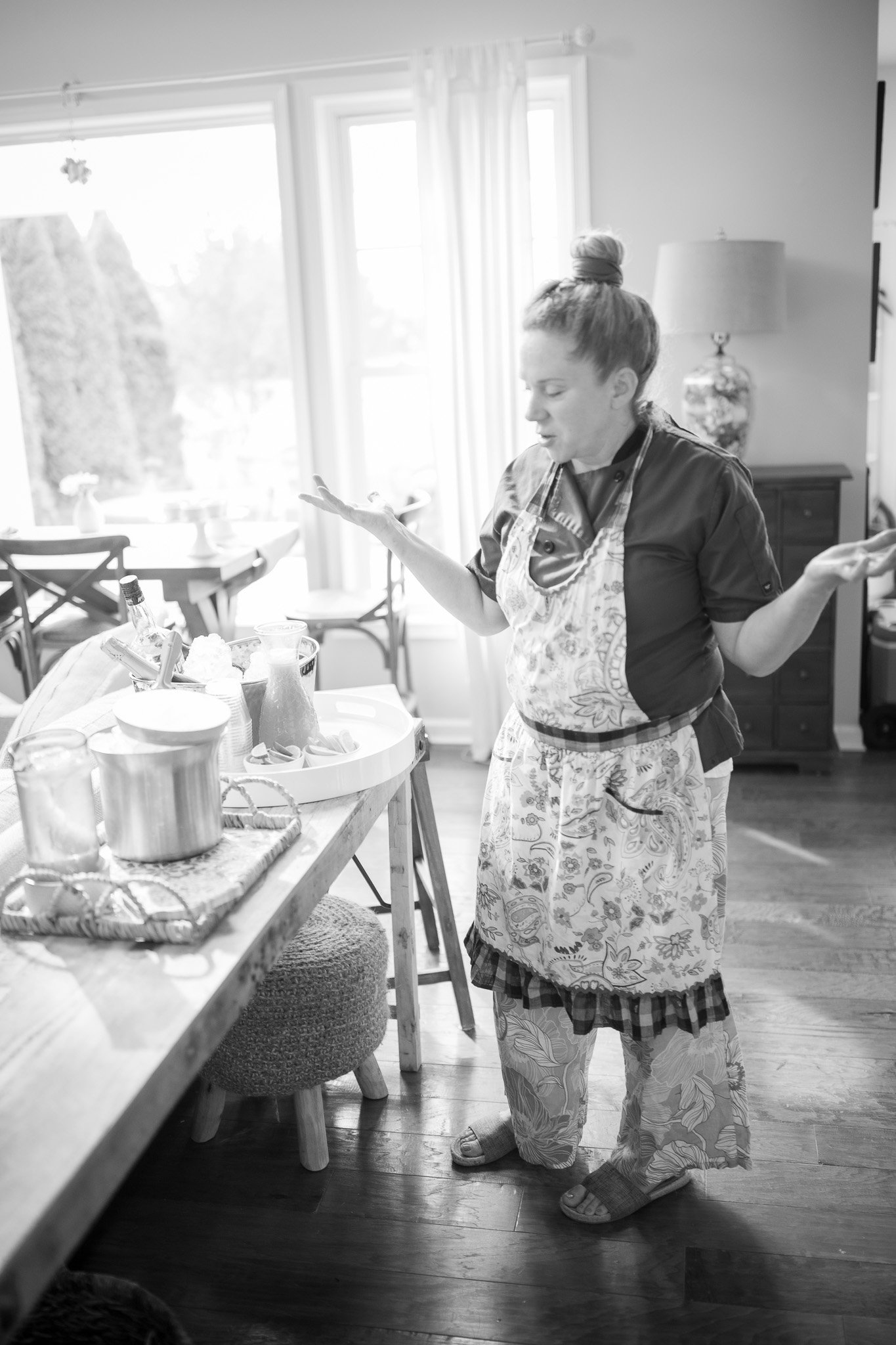 Person in kitchen wearing apron, standing near table with kitchen items, black and white photo.