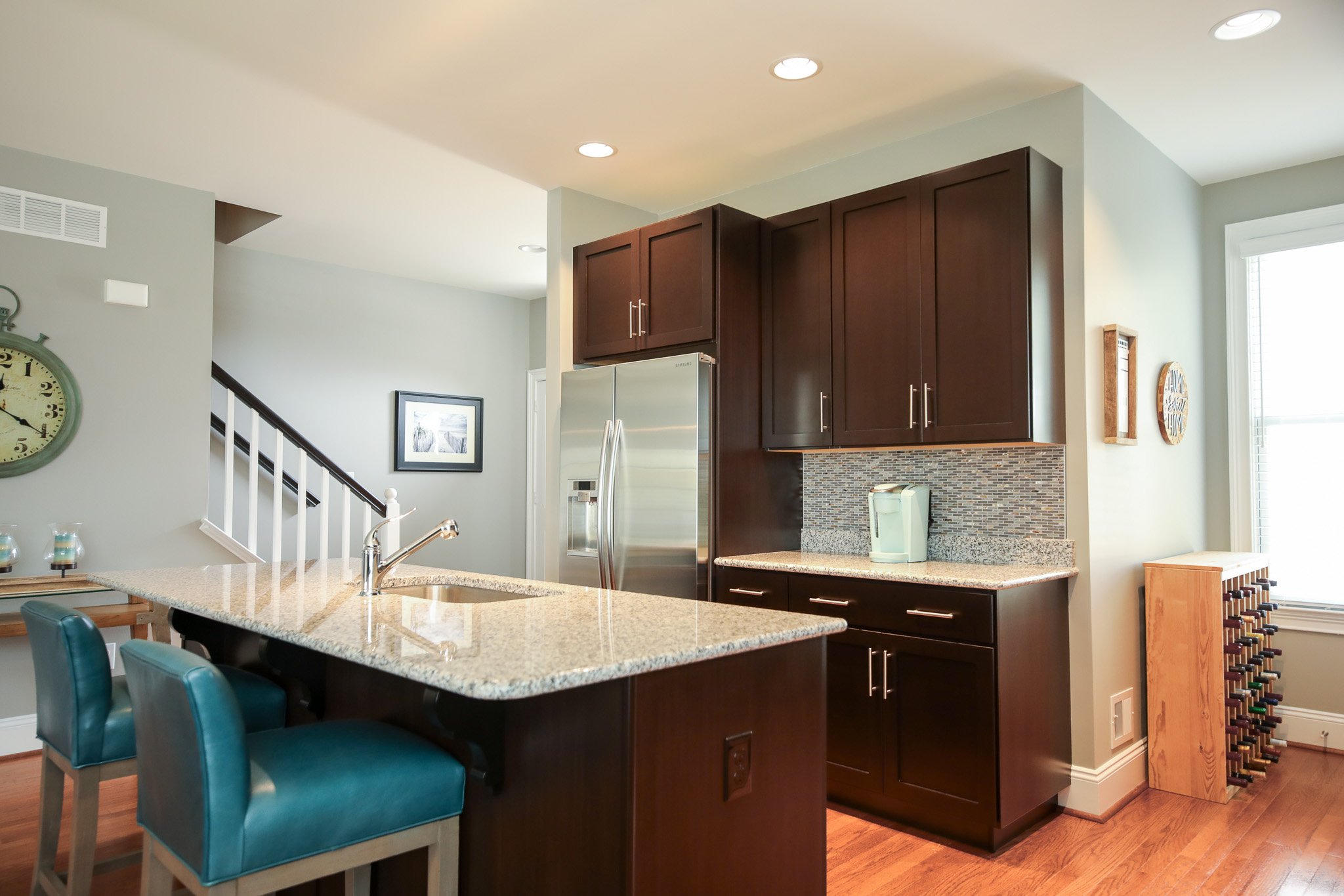 Modern kitchen with dark wood cabinets, stainless steel refrigerator, granite countertops, teal bar stools, coffee machine, and wine rack.