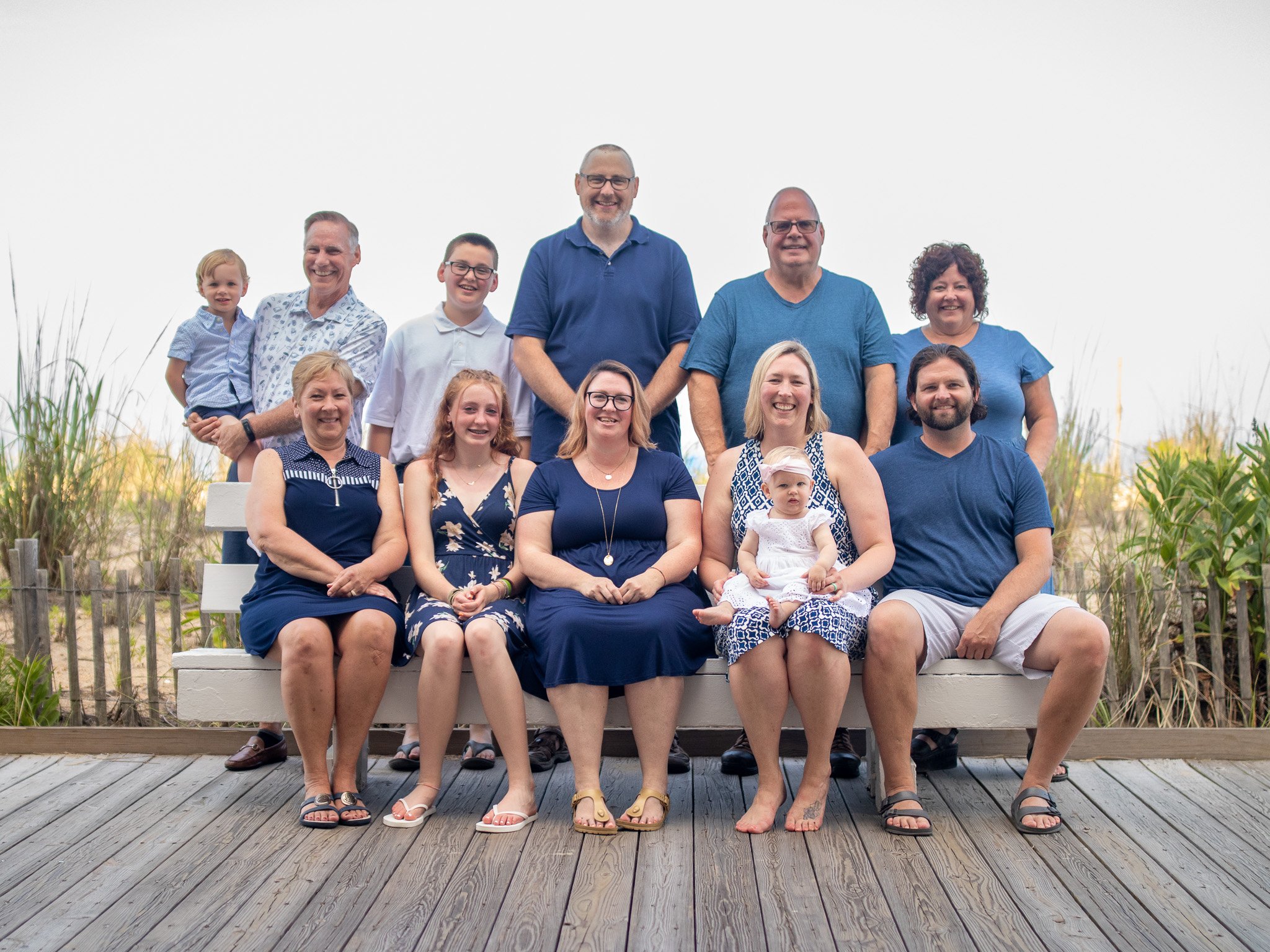 Group photo of several adults and children sitting and standing on a wooden deck by the beach, with a fence and dune grass in the background.