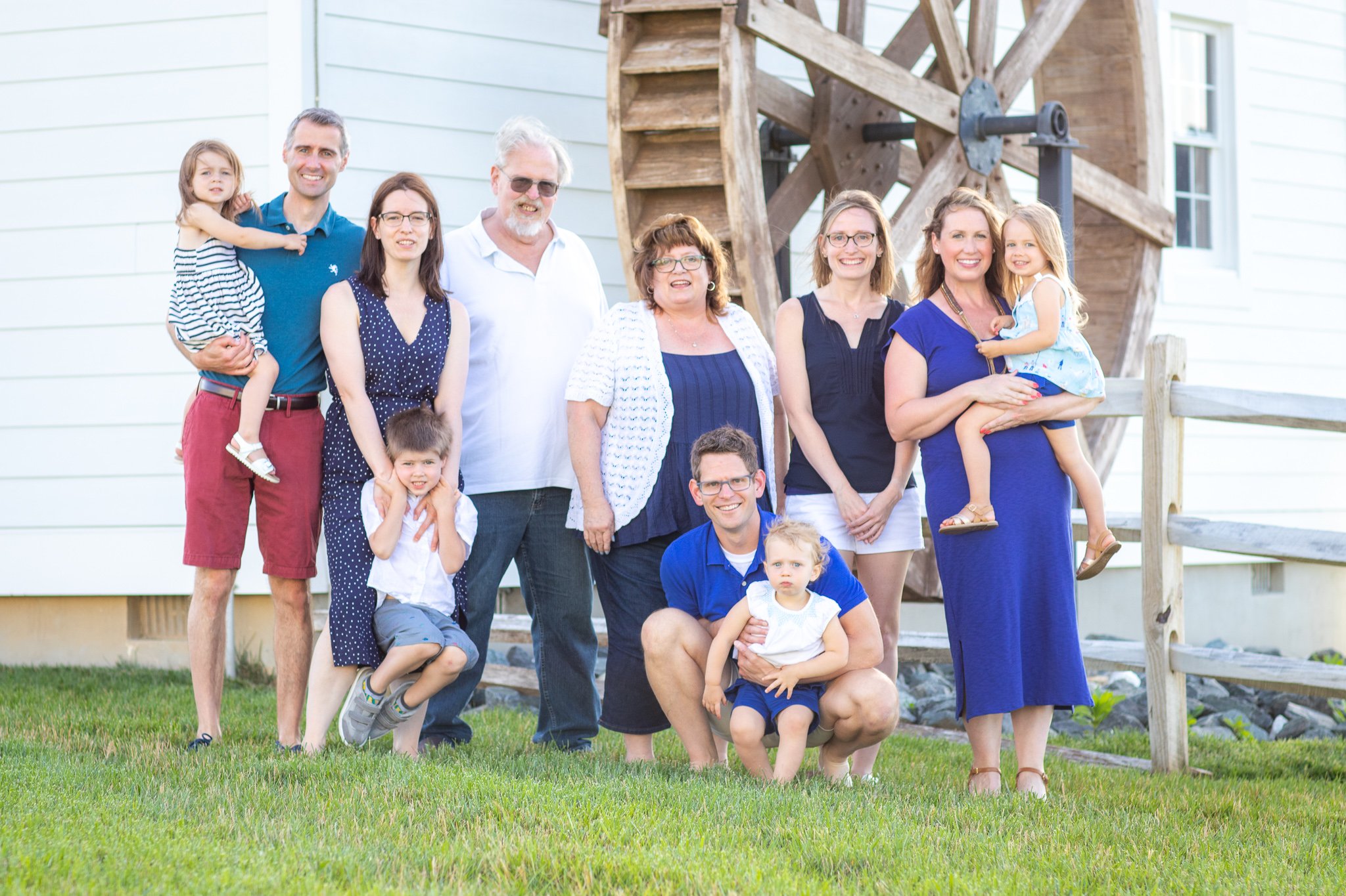 Family group photo in front of a wooden water wheel, with nine people including adults and children, outside on a grassy area.