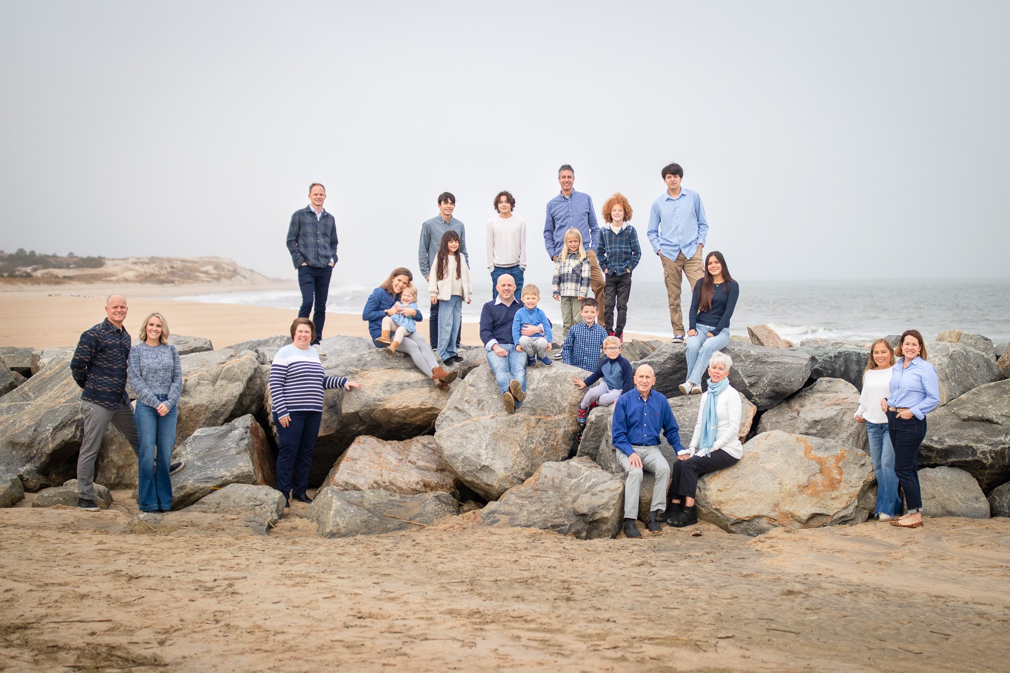 Large family group posing on rocks by the beach, cloudy day.