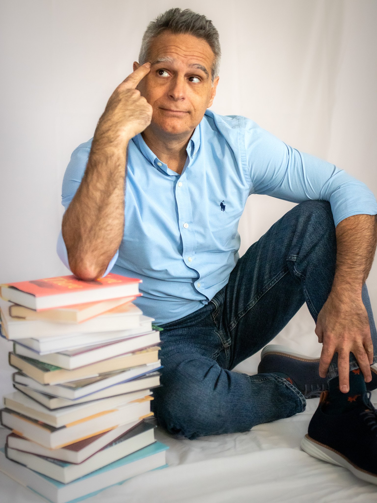 Man sitting next to a stack of books, wearing a blue shirt and jeans, with a thoughtful expression.