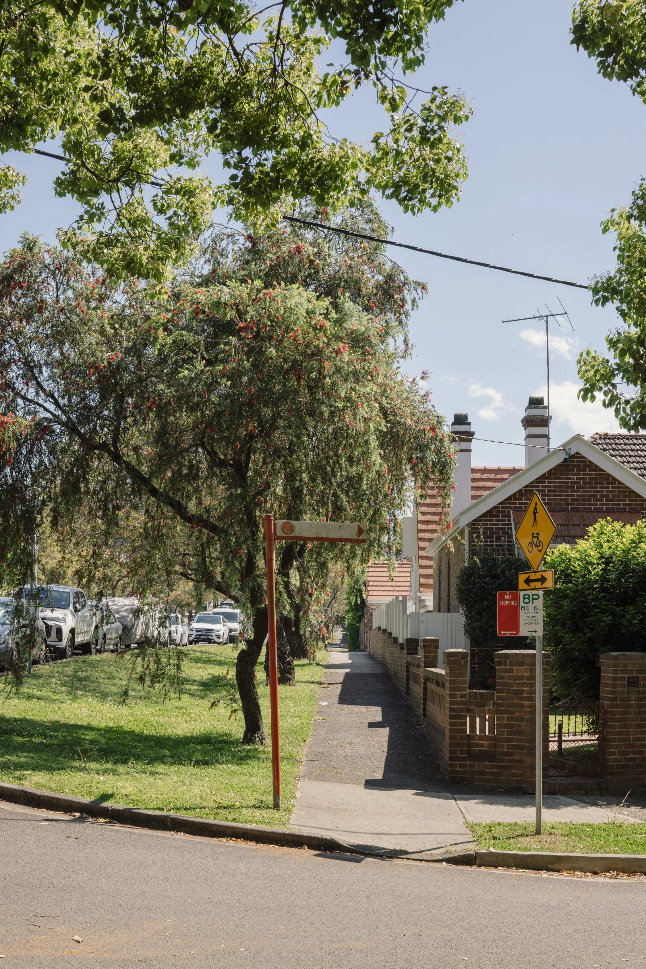 Sidewalk with street signs, brick house, trees, parked cars, and a clear sky.