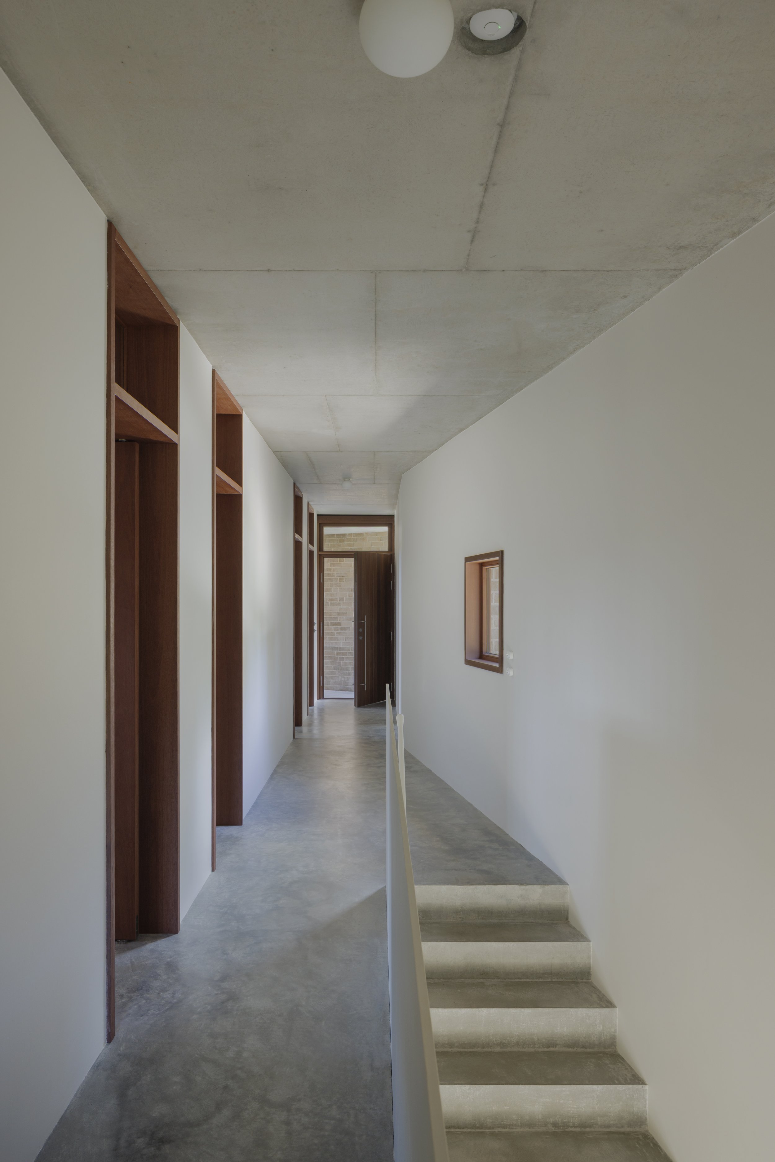 Modern interior hallway with white walls, wooden framed windows, and concrete ceiling and floors, leading to a wooden door at the end.