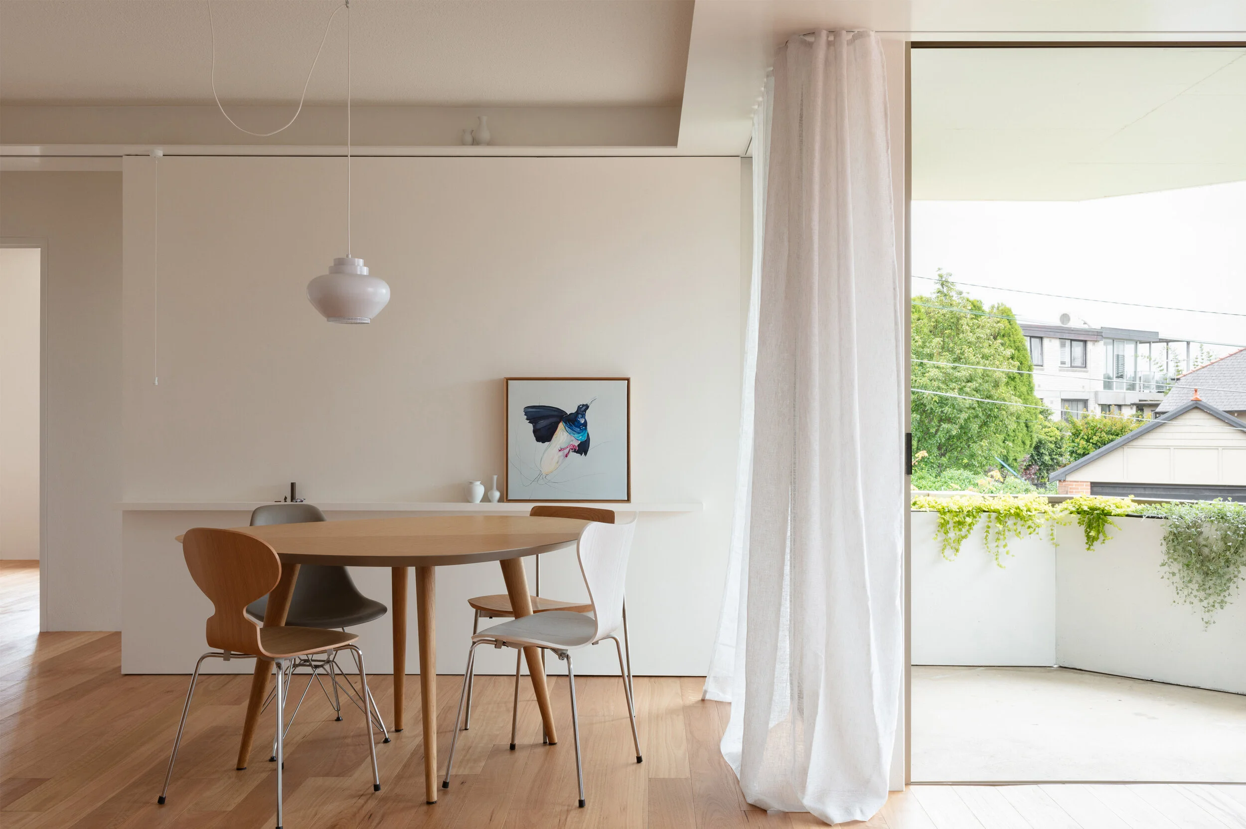 Minimalist dining area with a wooden table and four chairs, a large window with sheer curtains opening to a small balcony with greenery, and artwork of a butterfly on the wall.