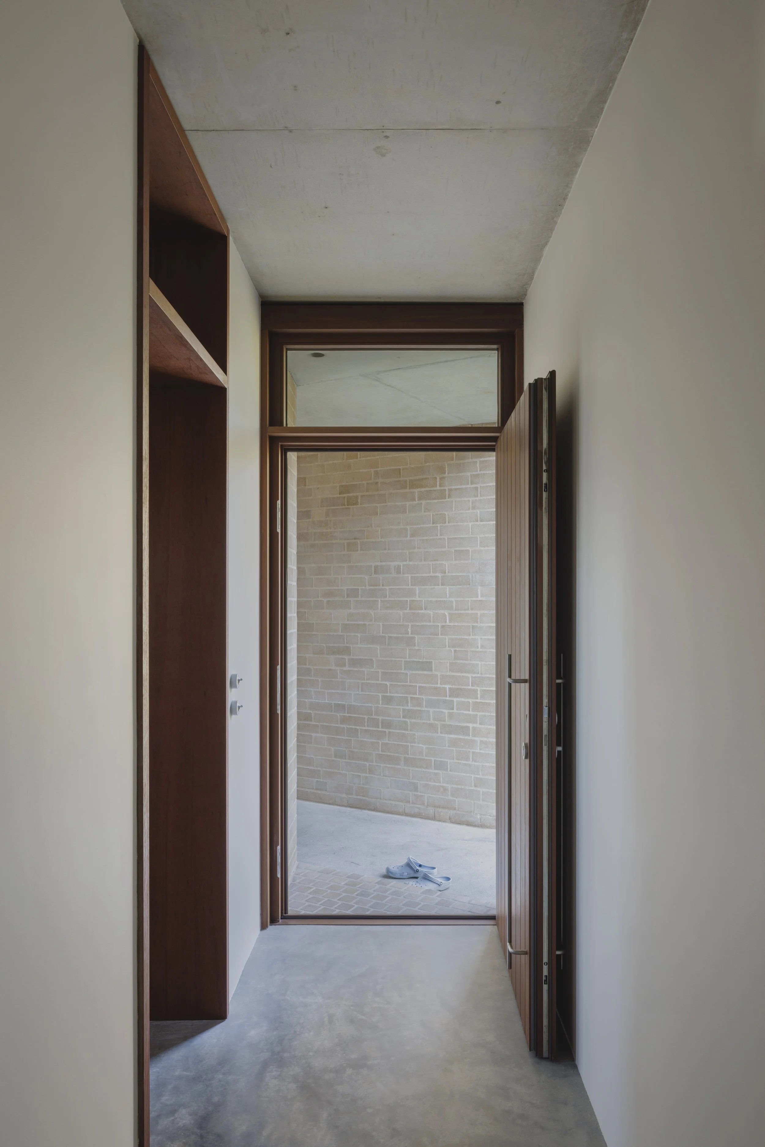Interior view of an entryway with a door open to a small patio or backyard, visible brick wall outside, and a pair of shoes on the ground. The hallway has plain walls, an unfinished concrete ceiling, and built-in wooden shelves on the left side.