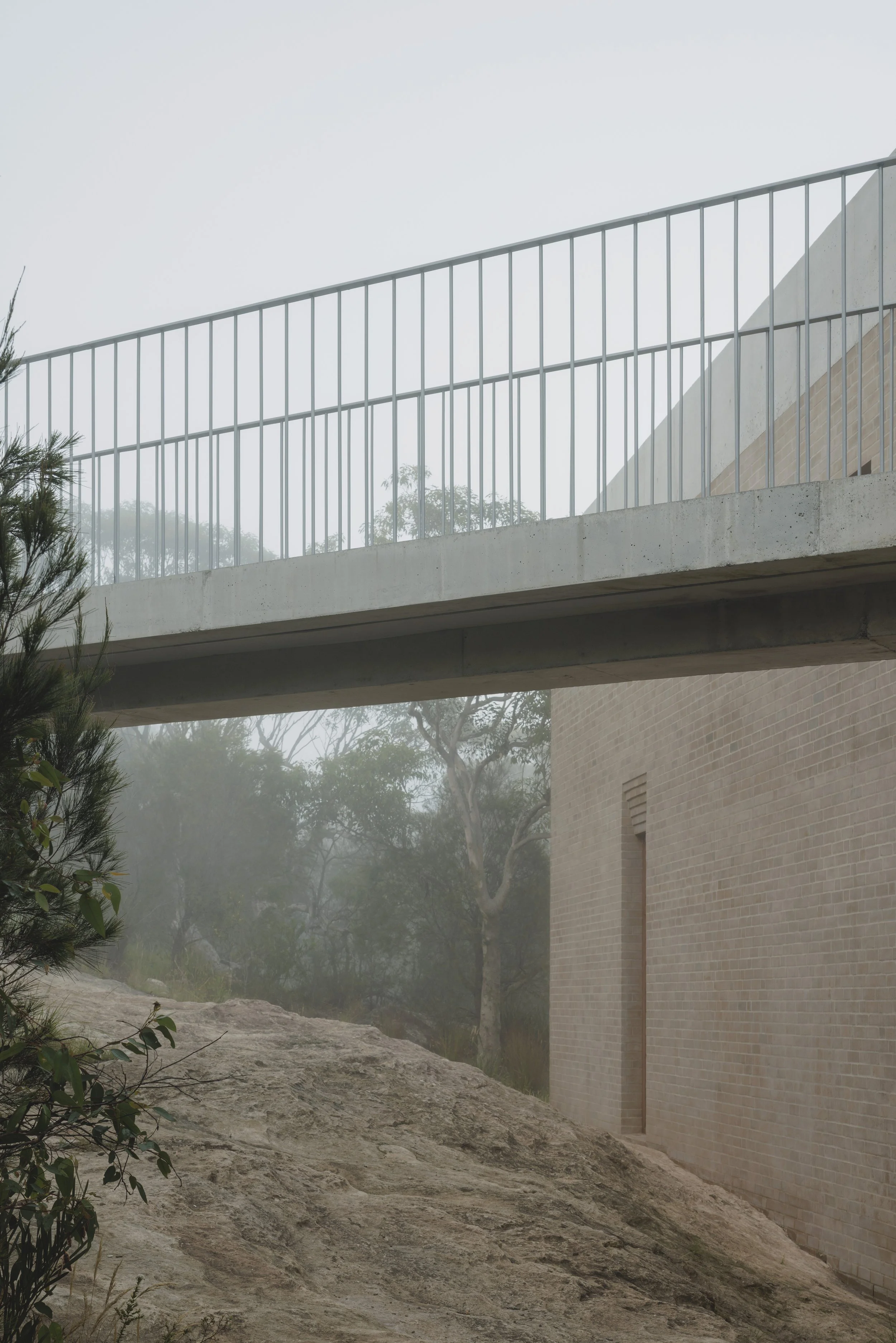 A foggy outdoor scene featuring a modern concrete and brick structure with a walkway and railing, trees, rocks, and fog in the background.