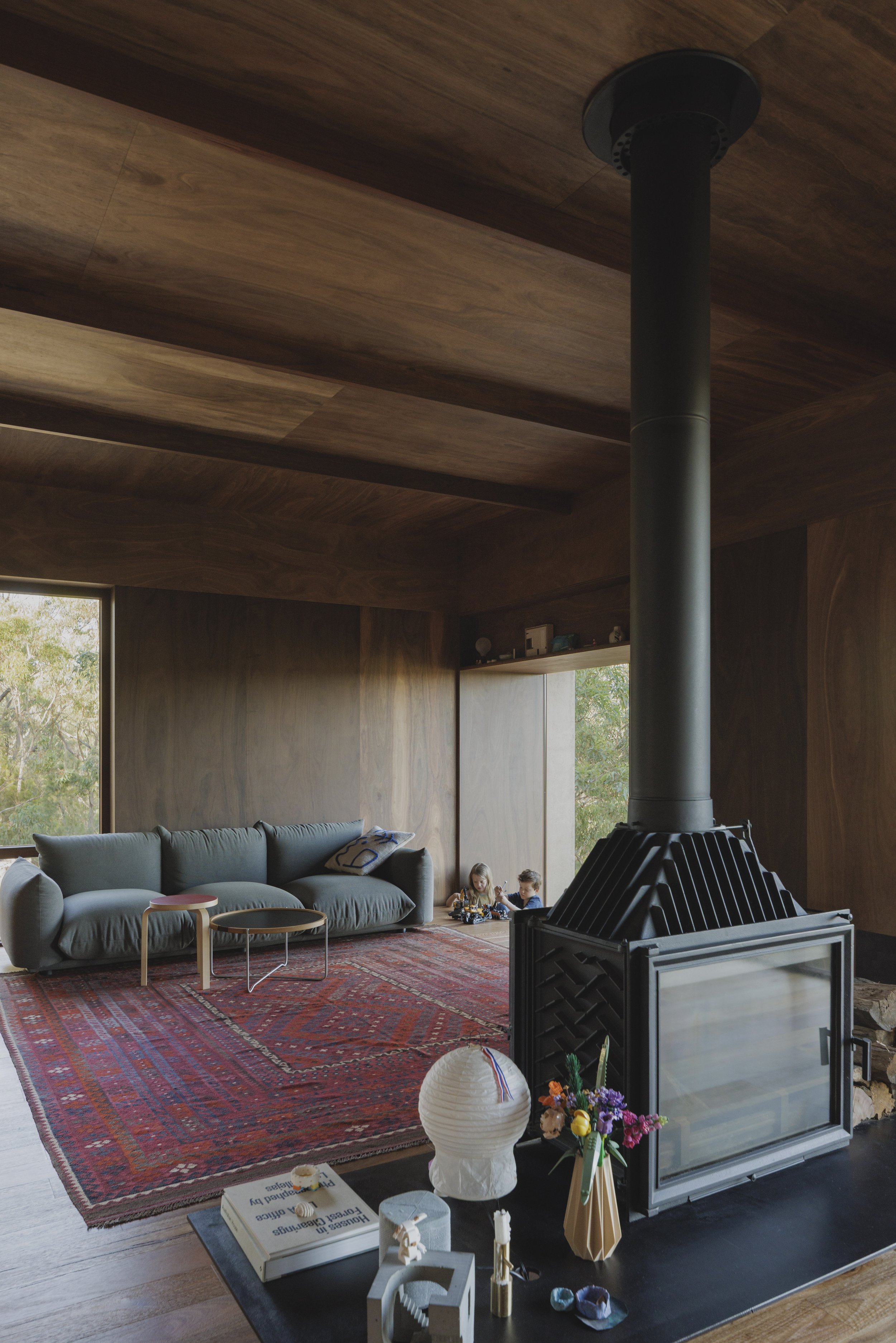 A living room with a large gray sofa, wooden walls, a patterned red rug, a modern wood-burning stove, and two children playing on the floor by a large window with trees outside.