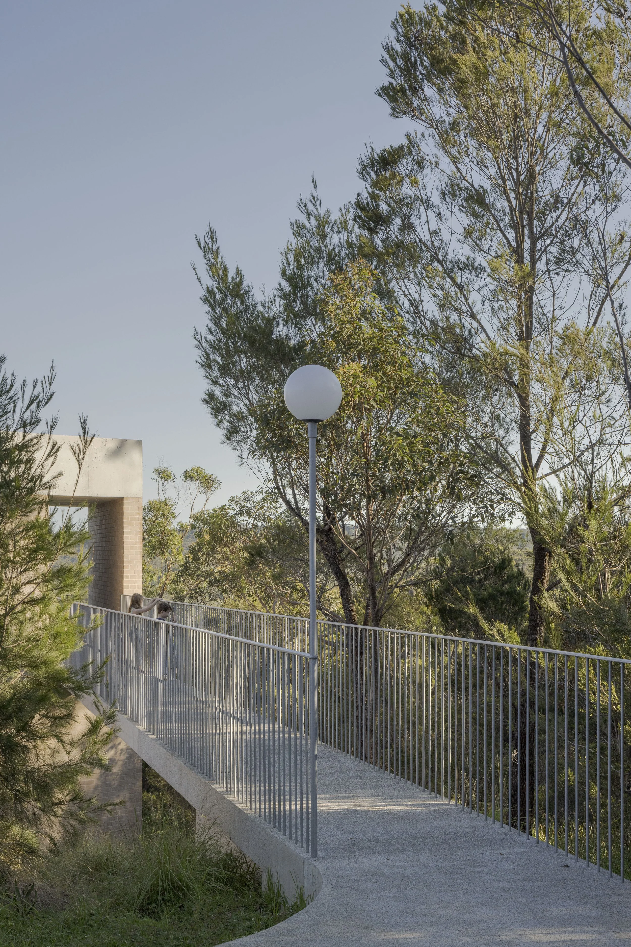 A concrete footpath with a metal railing curves through a green park with tall trees and shrubs. A street lamp stands along the path, with two children leaning over the railing and looking down.