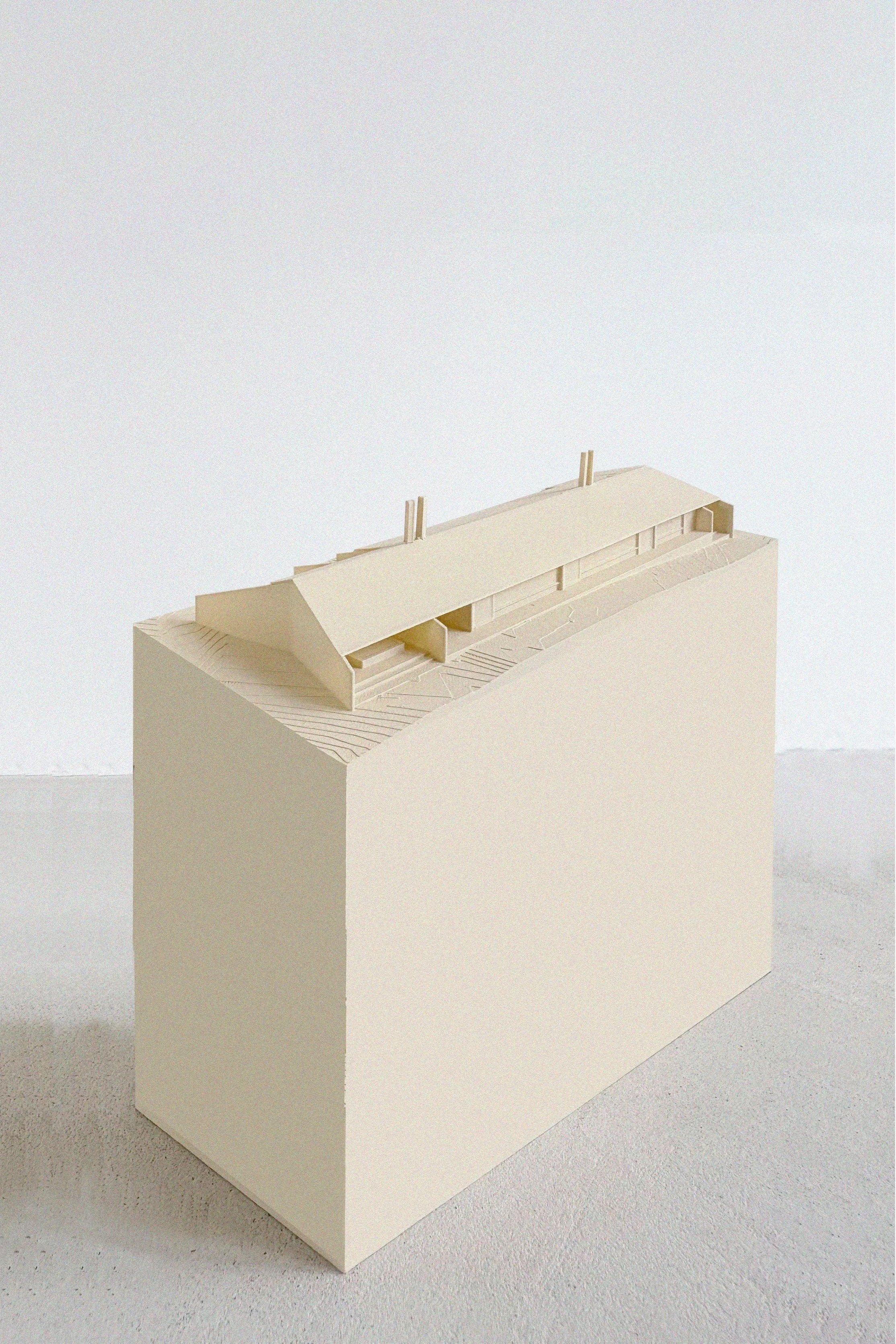 A small beige architectural model of a building on a rectangular stand, with a sloped roof and minimal design, placed on a light-colored surface against a plain white background.