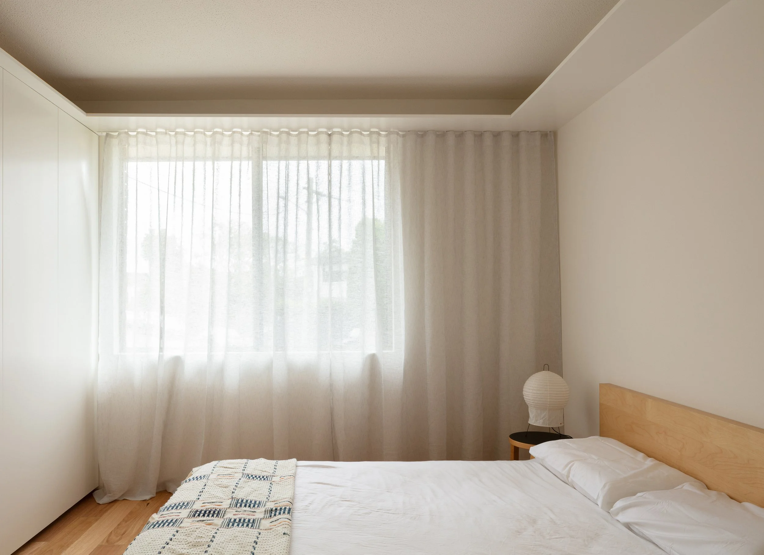 Simple bedroom with a bed, white bedding, a wooden headboard, a side table with a paper lantern lamp, and a window with sheer curtains letting in natural light.