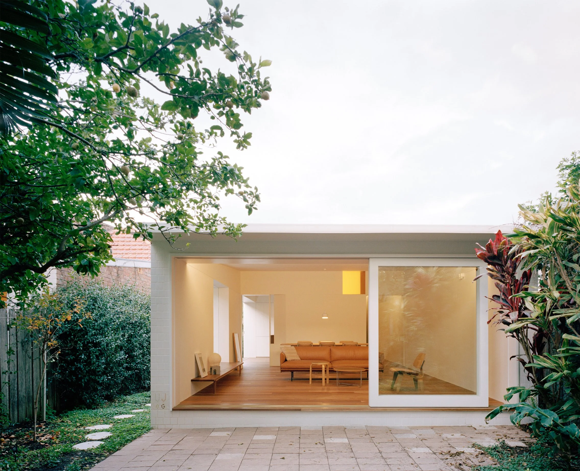 Modern single-story house with large sliding glass door open to backyard, visible interior with minimalist furniture and wooden floors, surrounded by trees and shrubs.