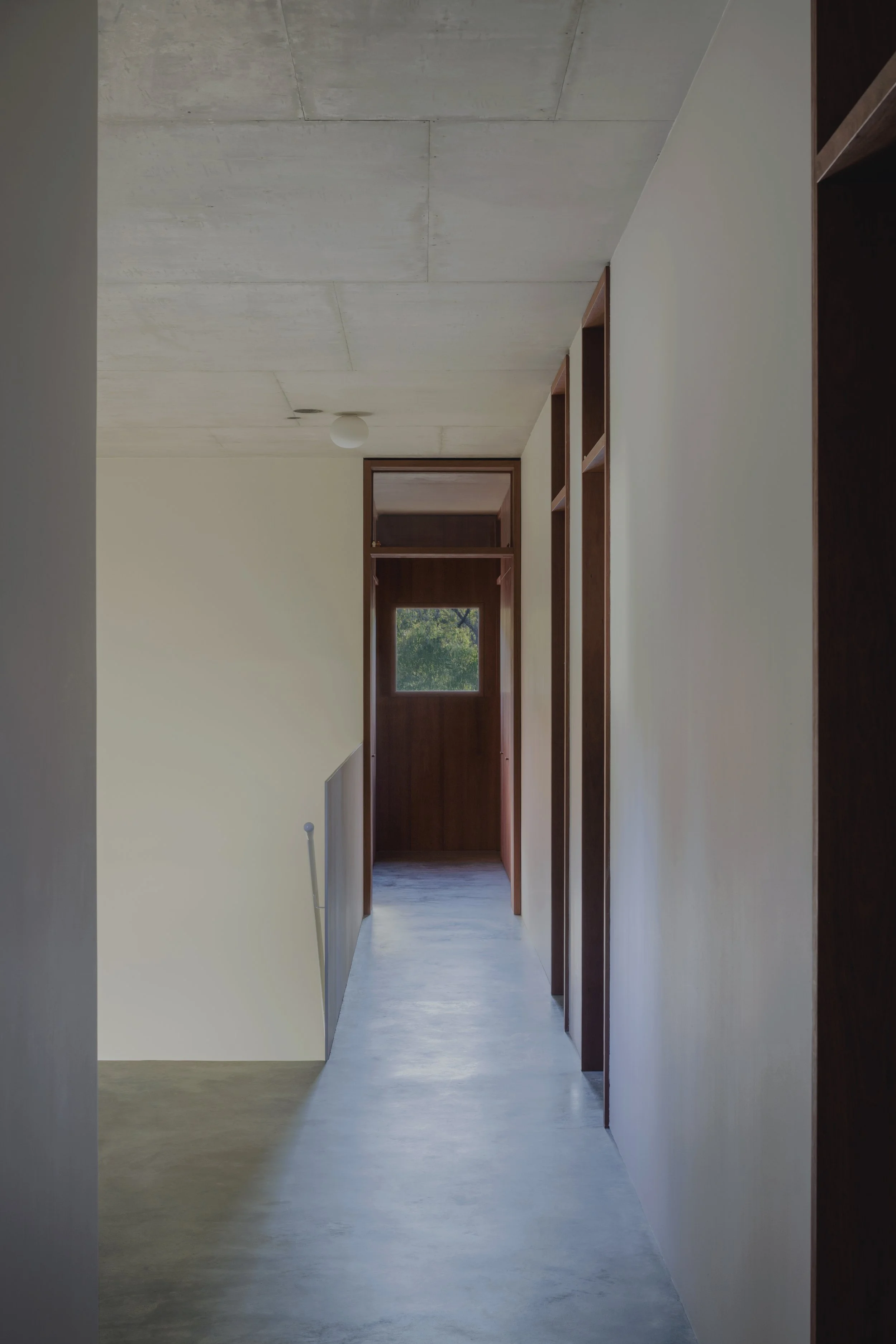 Empty hallway with a concrete ceiling, cream-colored walls, and dark wooden doorframes leading to small rooms. A window at the end shows greenery outside.