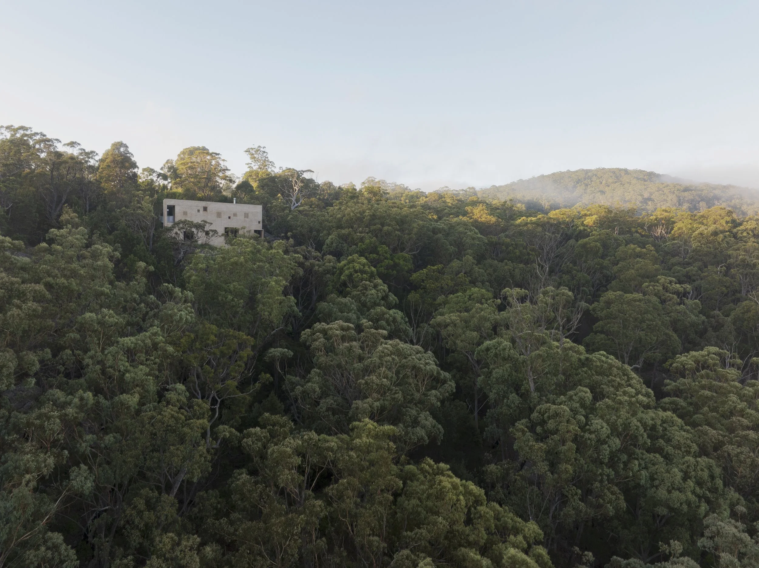 A modern, rectangular building with small windows, perched on a hillside surrounded by dense green forest, with mountains and a light sky in the background.